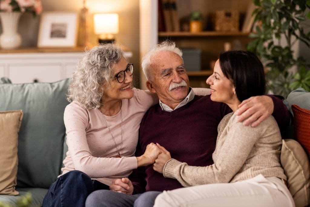 Smiling daughter hugging with her senior parents while she is visiting them, they sitting on couch in living room.